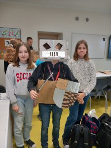 Uczeń pozuje do zdjęcia w wykonanej przez siebie zbroi. Na głowie ma hełm a w dłoniach trzyma tarczę i miecz. Po obu jego stronach stoją jego koleżanki z klasy.