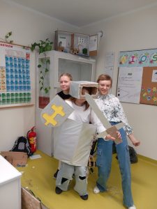 Uczennica pozuje do zdjęcia w wykonanej przez siebie zbroi rycerskiej. Dziewczynka trzyma w dłoniach tarczę i miecz a na głowie ma hełm. Za nią stoją dwie uczennice również pozujące do zdjęcia.