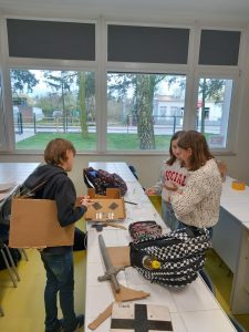 Troje uczniów podczas przygotowań do zaprezentowania projektu. Stoją przy długim stole, na którym leżą elementy przygotowywanej przez nich zbroi.