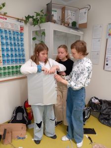 Dwie uczennice pomagają koleżance założyć przygotowaną przez nich zbroję rycerską.