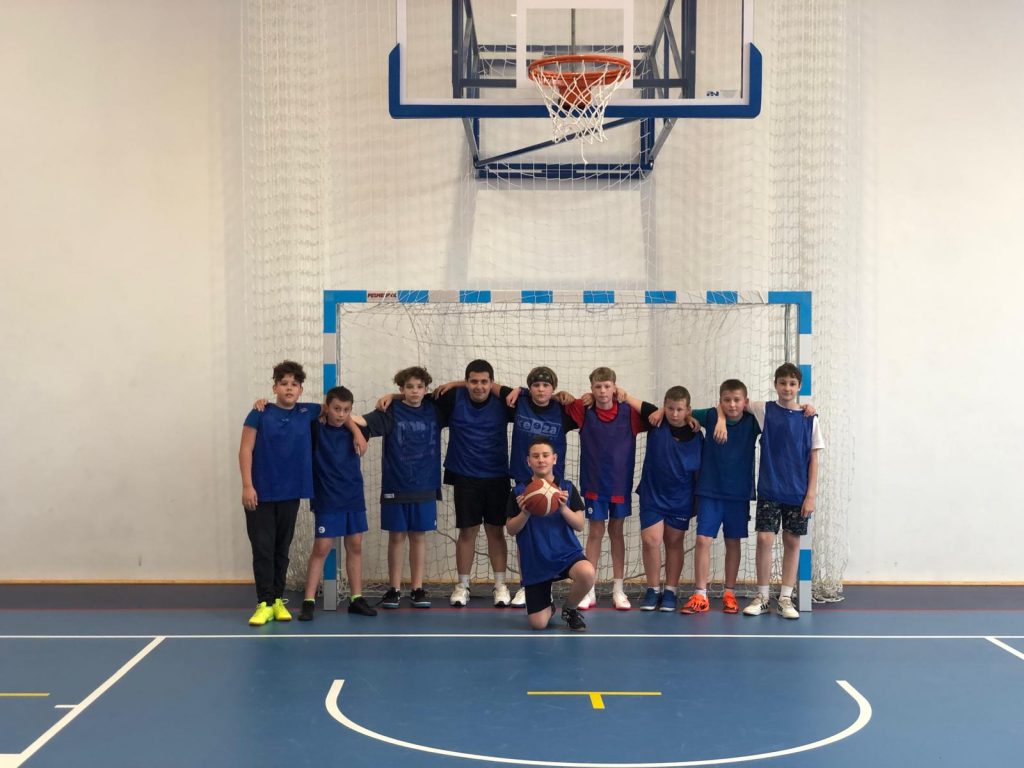 W świetle bramki na sali gimnastycznej do wspólnego zdjęcia pozują chłopcy reprezentujący w zawodach Szkołę Podstawową w Ostaszewie. Chłopcy ubrani są w niebieskie koszulki i stoją w jednym rzędzie opierając swoje ręce na ramionach kolegów stojących obok. Przed nimi na jednym kolanie kuca chłopiec trzymający przed sobą piłkę.