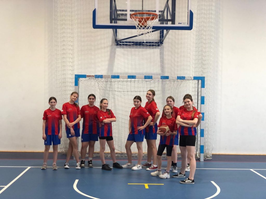 Uczennice reprezentujące Szkołę Podstawową w Ostaszewie stoją w świetle bramki na sali gimnastycznej. Dziewczynki mają czerwone koszulki i niebieskie lub czarne spodenki. Stoją luźno, część bokiem do zdjęcia. Jedna z nich trzyma piłkę.