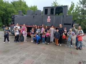 Uczniowie pozują do wspólnego zdjęcia wraz ze swoimi mamami. Wszyscy stoją przed dużą lokomotywą.