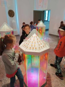 Uczniowie chodzą pomiędzy kolorowymi, szklanymi lampionami, z których wydobywa się kolorowe światło. Dzieci przyglądają się im z zaciekawieniem.