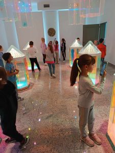 Uczniowie chodzą pomiędzy kolorowymi, szklanymi lampionami, z których wydobywa się kolorowe światło. Dzieci przyglądają się im z zaciekawieniem. Uczennica widoczna na pierwszym planie robi zdjęcie lampionowi stojącemu przed nią.