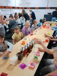 Uczniowie klasy pierwszej siedzą przy długim stole. Na stole stoją kubeczki z kolorowymi płynami oraz silikonowe foremki.