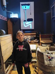 Uczennica pozuje do zdjęcia stojąc na tle schematu zawieszonego na ścianie za nią. Schemat przedstawia spłuczkę i spływającą wodę.