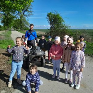 Uczniowie oddziału przedszkolnego pozują do wspólnego zdjęcia stojąc na drodze gruntowej. Pomiędzy uczniami stoi Pan trzymając taczkę na której umieszczone są worki z zebranymi przez dzieci śmieciami.
