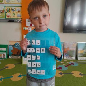 Uczeń oddziału przedszkolnego pozuje do zdjęcia trzymając przed sobą folię, pod która umieszczone są literki. Chłopiec ułożył z nich następujące wyrazy: Iga, Olek, Ala, koza, sok, mama.