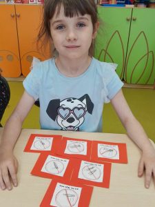 Uczennica siedzi przy stoliku z rękoma opartymi o niego. Przed dziewczynką leży 6 rysunków w czerwonych ramkach ułożonych w piramidę. Na rysunkach widoczne są podpisy i przekreślone przedmioty narysowane w czerwonych kółkach.