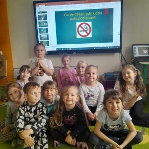 Uczniowie oddziału przedszkolnego pozują do wspólnego zdjęcia. Dzieci siedzą na dywanie a za nimi na ekranie wyświetla się slajd z napisem "Co się dzieje, gdy ludzie palą papierosy?". Pod napisem widoczna jest grafika przekreślonego papierosa w czerwonym kółku.