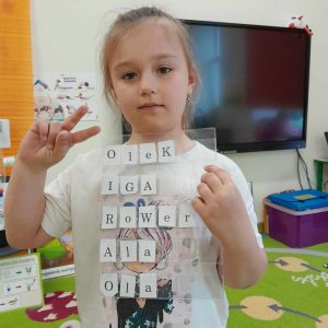 Uczennica oddziału przedszkolnego pozuje do zdjęcia trzymając jedna dłonią folię, pod która umieszczone są literki. W drugiej ręce dziewczynka ma wyprostowane dwa palce i trzyma je na wysokości głowy. Dziewczynka ułożyła z nich następujące wyrazy: Olek, Iga, rower, Ala, Ola.
