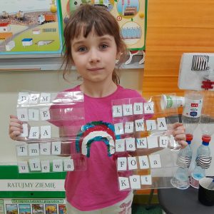 Uczennica oddziału przedszkolnego pozuje do zdjęcia trzymając przed sobą dwie folie, pod którymi umieszczone są literki. Dziewczynka ułożyła z nich następujące wyrazy: dom, ale, Aga, Tmek, ulewa, Kuba, Jiwja, soku, foka, kot.