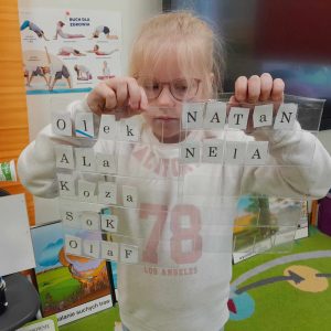 Uczennica oddziału przedszkolnego pozuje do zdjęcia trzymając przed sobą dwie folie, pod którymi umieszczone są literki. Dziewczynka ułożyła z nich następujące wyrazy: Olek, Ala, koza, sok, Olaf, Natan, Nela.