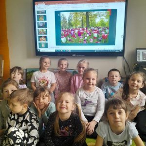 Uczniowie oddziału przedszkolnego pozują do wspólnego zdjęcia. Dzieci siedzą na dywanie. Za nimi na tablicy wyświetla się slajd ze zdjęcie ukwieconej łąki.
