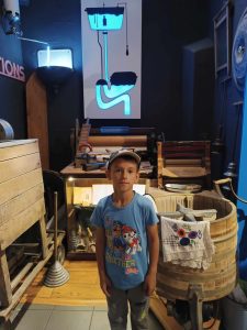 Uczeń pozuje do zdjęcia stojąc na tle schematu zawieszonego na ścianie za nim. Schemat przedstawia spłuczkę i spływającą wodę.