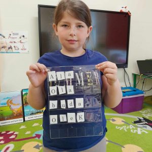 Uczennica oddziału przedszkolnego pozuje do zdjęcia trzymając przed sobą folię, pod która umieszczone są literki. Dziewczynka ułożyła z nich następujące wyrazy: Ala, Ola, buda, Olek.