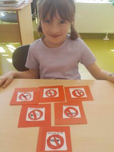 Uczennica siedzi przy stoliku z rękoma opartymi o niego, uśmiecha się. Przed dziewczynką leży 6 rysunków w czerwonych ramkach ułożonych w piramidę. Na rysunkach widoczne są podpisy i przekreślone przedmioty narysowane w czerwonych kółkach.