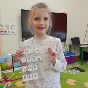 Uczennica oddziału przedszkolnego pozuje do zdjęcia trzymając przed sobą folię, pod która umieszczone są literki. Dziewczynka ułożyła z nich następujące wyrazy: Ala, Olek, buda, sok, but.
