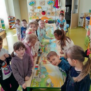 Uczniowie oddziału przedszkolnego stoją przy długim stole, na którym rozłożona jest kolorowa plansza. Część z dzieci patrzy się w stronę obiektywu aparatu.