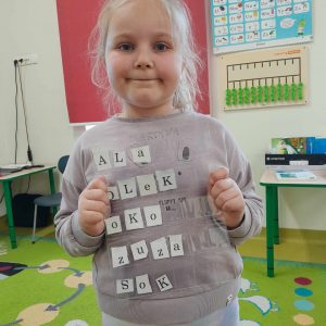 Uczennica oddziału przedszkolnego pozuje do zdjęcia trzymając przed sobą folię, pod która umieszczone są literki. Dziewczynka ułożyła z nich następujące wyrazy: Ala, Olek, oko, Zuza, sok.
