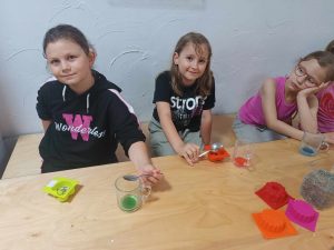 Trzy dziewczynki siedzą przy wspólnym stole. Jedna z nich podpiera głowę ręką opartą na stole. Dwie pozostałe dziewczynki trzymają w dłoniach łyżeczki i uśmiechają się do obiektywu.