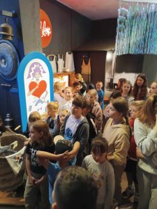 Uczniowie klasy pierwszej stoją wspólnie w jednej z sali zwiedzanego obiektu. Dzieci patrzą przed siebie.