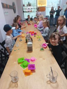 Uczniowie klasy pierwszej siedzą przy długim stole. Na stole stoją kubeczki z kolorowymi płynami oraz silikonowe foremki.