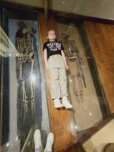 Dziewczynka leży na podłodze. Obok niej za przeszkloną szybą leżą dwa ludzkie szkielety.