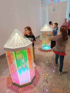 Uczniowie chodzą pomiędzy kolorowymi, szklanymi lampionami, z których wydobywa się kolorowe światło.