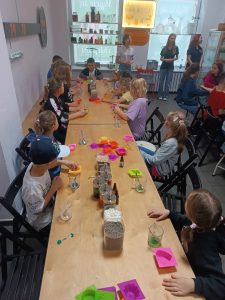 Uczniowie klasy pierwszej siedzą przy długim stole. Na stole stoją kubeczki z kolorowymi płynami oraz silikonowe foremki.