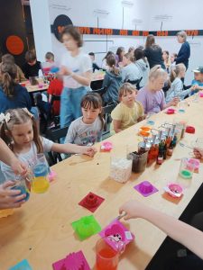 Uczniowie klasy pierwszej siedzą przy długim stole. Na stole stoją kubeczki z kolorowymi płynami oraz silikonowe foremki.