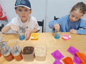 Dwoje dzieci siedzi przy stole. Uczniowie mieszają łyżeczkami niebieski płyn umieszczone w stojących przed nimi szklankami.