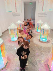 Uczniowie klasy pierwszej stoją w jednym rzędzie naprzemiennie wychylając głowy w przeciwne strony. Po ich lewej i prawej stronie ustawione są duże przeszklone lampiony, z których wydobywa się kolorowe światło.