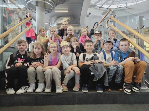 Uczniowie klasy pierwszej pozują do wspólnego zdjęcia wraz ze swoją wychowawczynią. Dzieci siedzą na stopniach szerokich schodów.