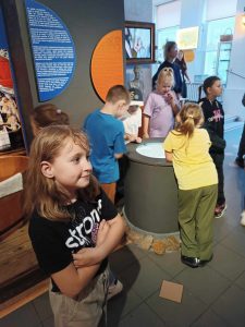 Uczennica widoczna na pierwszym planie stoi ze skrzyżowanymi rękami patrząc się przed siebie. W tle widoczni są uczniowie stojący wokół beczki z której wydobywa się jasne światło.