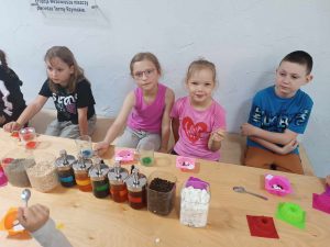 Czworo uczniów klasy pierwszej siedzi przy długim stole. Na stole stoją kubeczki z kolorowymi płynami oraz silikonowe foremki. Przed dziećmi stoją w rzędzie dozowniki z kolorowymi płynami.