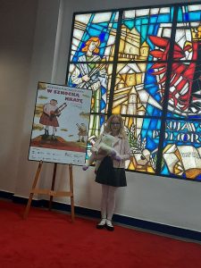 Uczennica nagrodzona w konkursie stoi trzymając w dłoni pluszową owieczkę, obok plakatu reklamującego konkurs z hasłem "W SZKOCKĄ KRATĘ". W tle widoczny jest duży witraż.