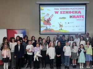Dzieci nagrodzone w konkursie pozują do wspólnego zdjęcia. W tle za nimi wyświetla się na rzutniku grafika dwóch szkotów stojących z dudami na górkach i napis: "FINAŁ KONKURSU PLASTYCZNEGO W SZKOCKĄ KRATĘ TORUŃ 2024". Poniżej słabo umieszczone są logo patronów i sponsorów, które są słabo widoczne.