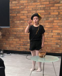 Uczennica podczas występu na konkursie. Dziewczynka ubrana jest na czarno i ma na twarzy wymalowane wąsy. Jedną rękę podnosi w górę, rozstawiając szeroko palce.