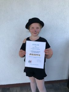 Uczennica stoi na tle białej ściany trzymając przed sobą otrzymany dyplom. Dziewczynka uśmiecha się, ubrana jest w czarny strój i kapelusz, a na twarzy ma wymalowane wąsy kota.