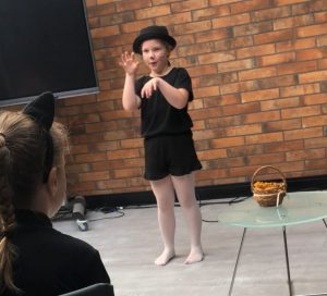 Uczennica podczas występu na konkursie. Dziewczynka stoi na scenie trzymając przed twarzą dłonie naśladujące ruchy kota.