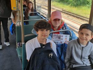 Troje uczniów pozuje do wspólnego zdjęcia siedząc w tramwaju. Jedna z dziewczynek trzyma mapkę.; W tle widać siedzących innych uczniów.