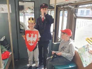 Uczeń pozuje do zdjęcia wraz z przewodnikiem. Chłopiec na głowie ma złotą koronę, a w dłoni trzyma czerwoną tarczę z białym orłem. Przewodnik ubrany jest w strój konduktora i trzyma przy ustach mikrofon. Obok siedzi innych uczeń spoglądający w ich stronę.