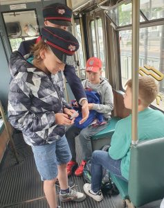 Uczeń w czapce konduktora kasuje bilety uczniom siedzącym w tramwaju. Obok niego stoi przewodnik w stroju konduktora wyciągający w jego stronę dłoń.