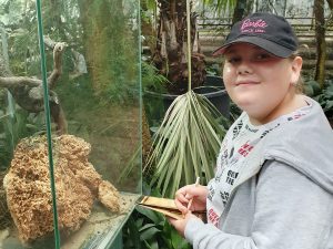 Uczennica stoi na przeciwko akwarium w którym jest umieszczone mrowisko. Dziewczynka patrzy w stronę aparatu. W dłoni trzyma kartę z zadaniami, którą wypełnia ołówkiem.