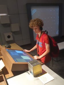 Uczeń ze słuchawkami na uszach stoi na przeciwko monitora interaktywnego, któremu się przygląda. W tle widać ekran zawieszony na ścianie.