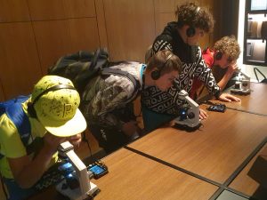 Czworo uczniów stoi przy długim stole. Troje z chłopców pochyla się nad mikroskopami ustawionymi przed nimi na stole. Wszyscy chłopcy mają na uszach słuchawki.