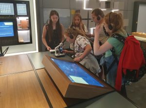 Pięć uczennic stoi w grupce przy stole, na którym ustawiony jest ekran i mikroskop. Jedna z dziewczynek pochyla się nad mikroskopem. Wszystkie dziewczynki mają na uszach słuchawki.