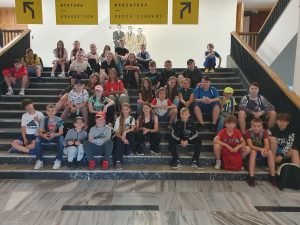 Uczniowie biorący udział w wycieczce siedzą na schodach i pozując do wspólnego zdjęcia. Za nimi widać makietę trzech naukowców a nad nimi zawieszono instrukcje ze strzałkami w lewo i w prawo oraz napisami: "WYSTAWA EXHIBITION", "MEDIATEKA MEDIA LIBRARY"